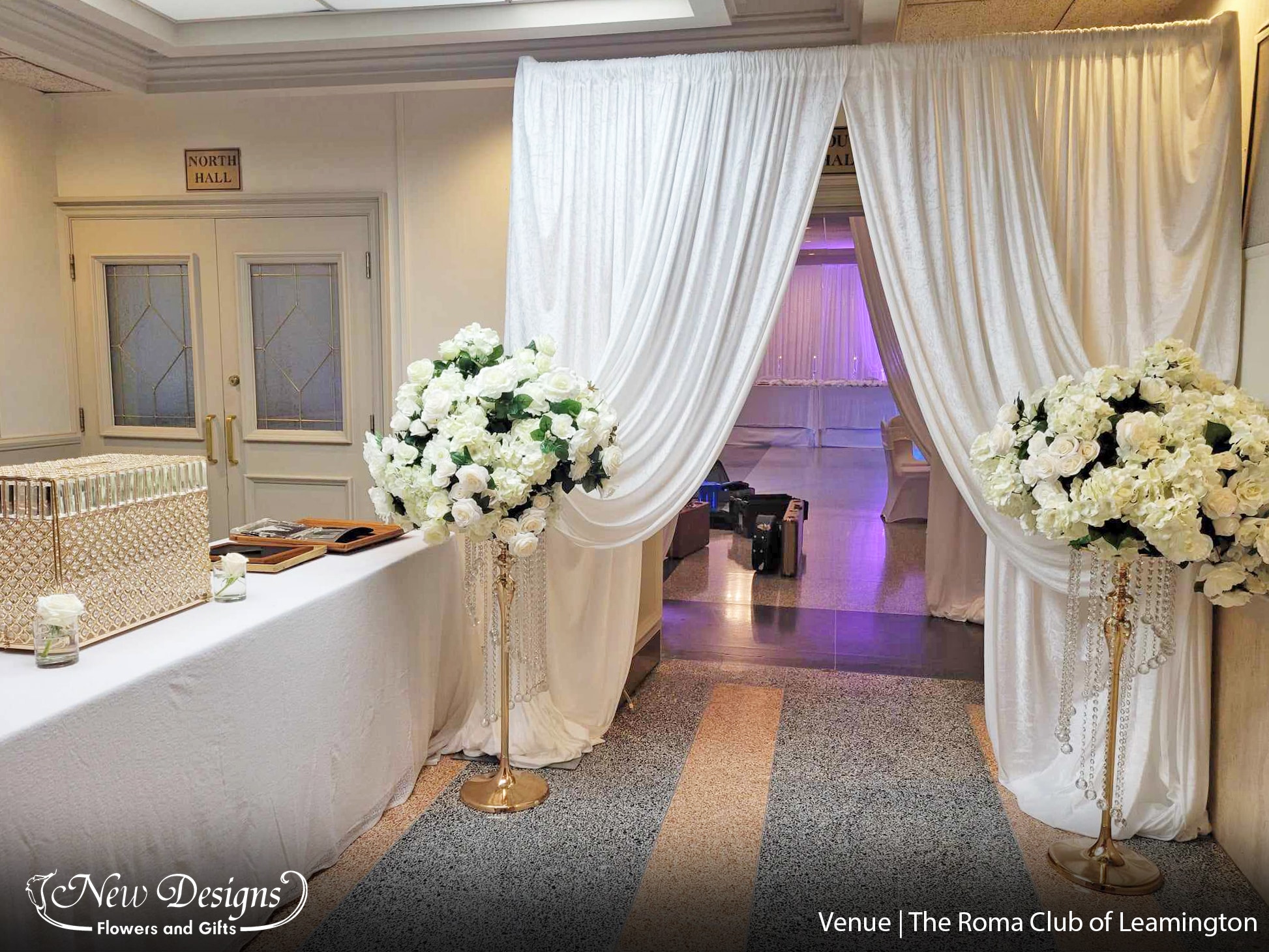 A draped entry way of white chiffon material. Adorned with 2 tall floor centerpieces, atop gold posts, with Jewels trailing down at gradually decreasing spirals. The arrangements full and lush with white hydrangeas roses and orchids.